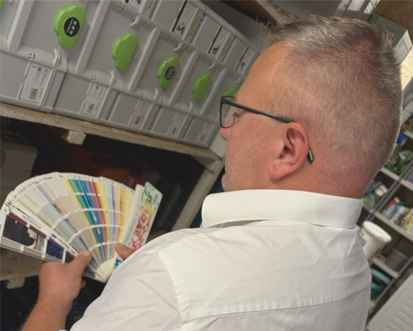 Eine Person mit kurzen Haaren und Brille, die ein weißes Hemd trägt, hält ein Farbmusterheft in der Hand und betrachtet beschriftete Behälter in Regalen in einem Raum, der wie ein Arbeits- oder Lagerbereich aussieht.